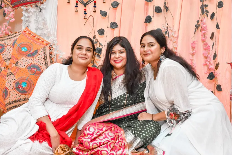 Three smiling Indian women in white kurtas and a green silk saree at a festive gathering