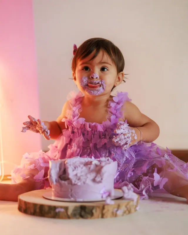 Toddler in a lilac butterfly dress with frosting on her face during a cake smash
