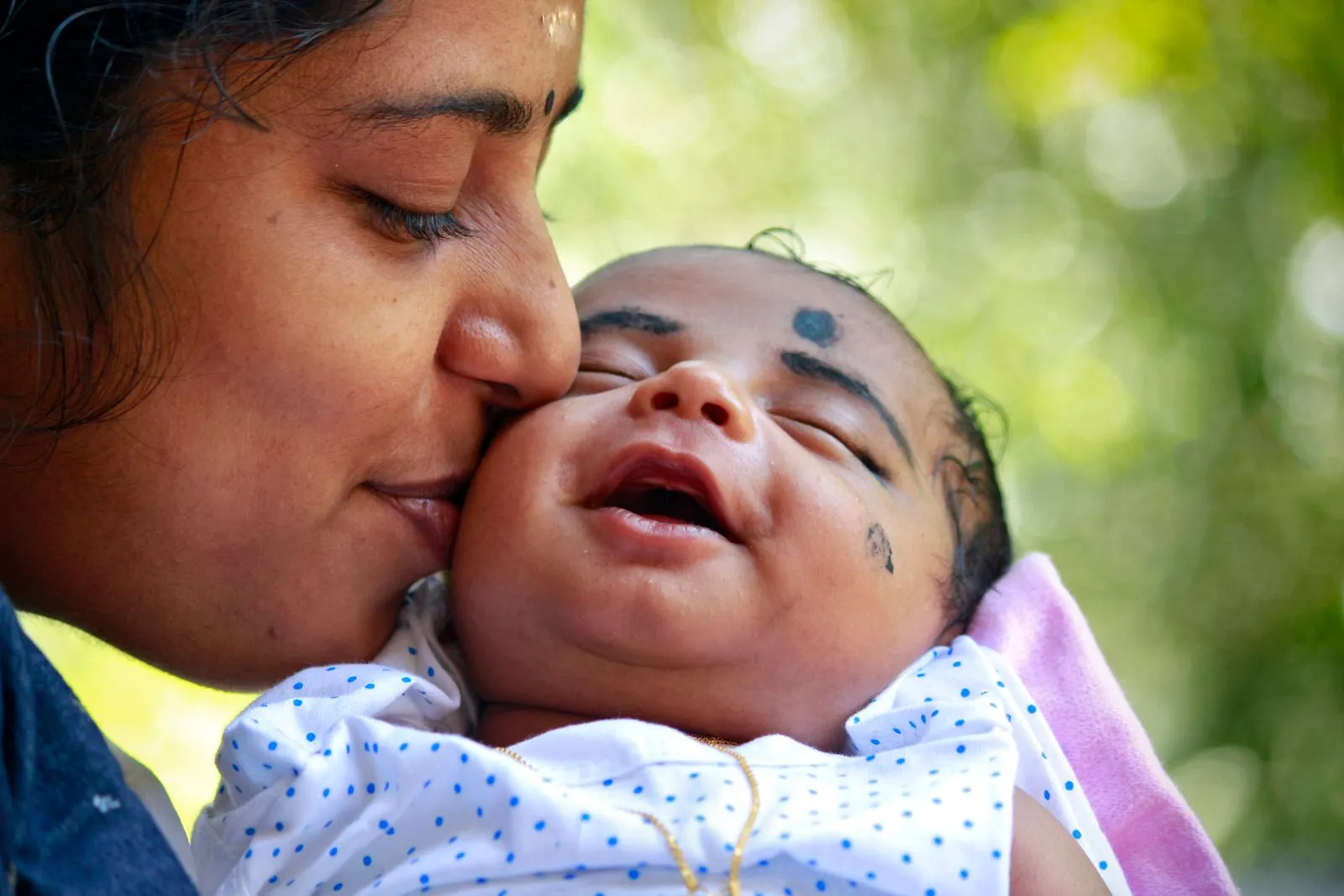 Indian mother tenderly kissing the cheek of her smiling newborn in a polka-dot outfit